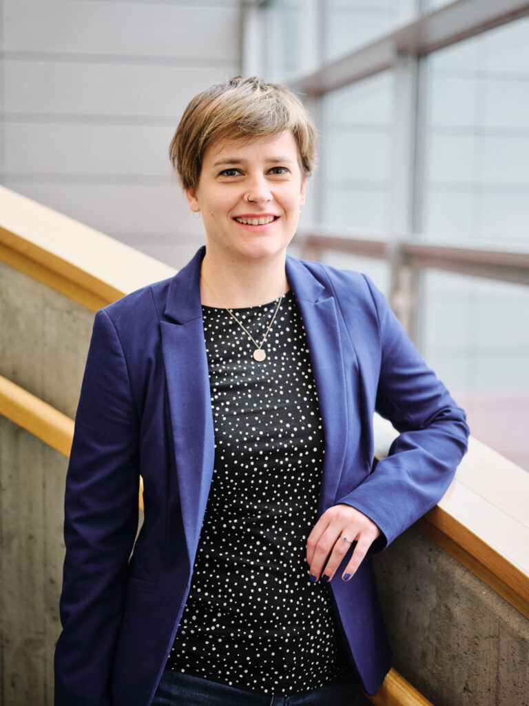 Lyhythiuksinen henkilö, jolla on sininen bleiseri ja musta paita, jossa on valkoisia pilkkuja, seisoo sisätiloissa kaiteen vieressä ja hymyilee kameralle.