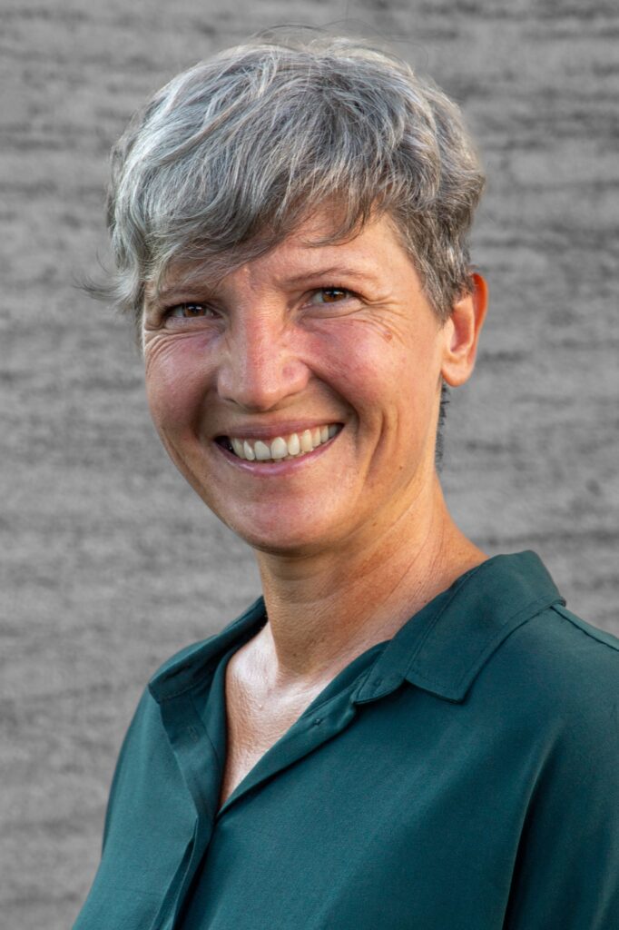 A person with short gray hair is smiling at the camera, wearing a dark green collared shirt, standing in front of a blurred gray textured background.