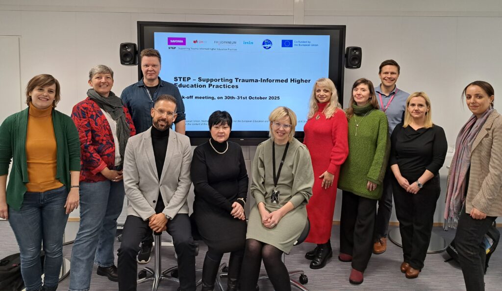 Kaksitoista ihmistä seisoo ja istuu hymyillen kokoushuoneessa suuren näytön edessä, jossa lukee "STEP - Supporting Trauma-Informed Higher Education Practices". Näytöllä näkyy päivämäärä ja hankekumppaneiden logot.
