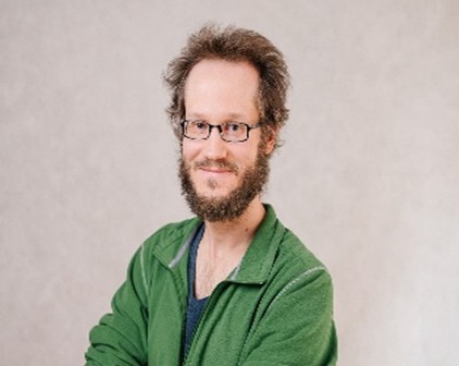 A man with glasses and a full beard, wearing a green jacket over a dark shirt, poses in front of a light gray background, smiling slightly.