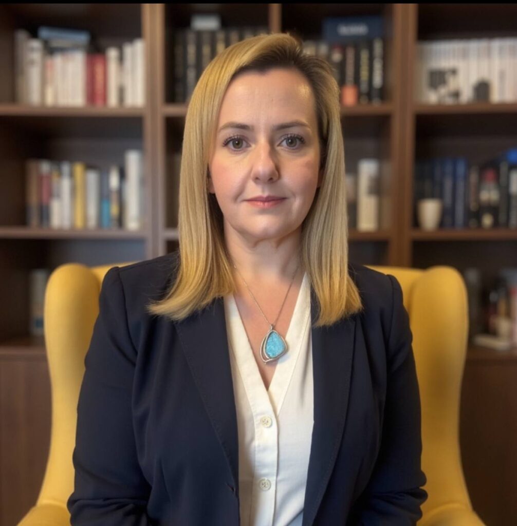 A woman with straight blonde hair, wearing a dark blazer and a white blouse with a turquoise pendant necklace, sits in a yellow chair in front of a bookcase filled with books.
