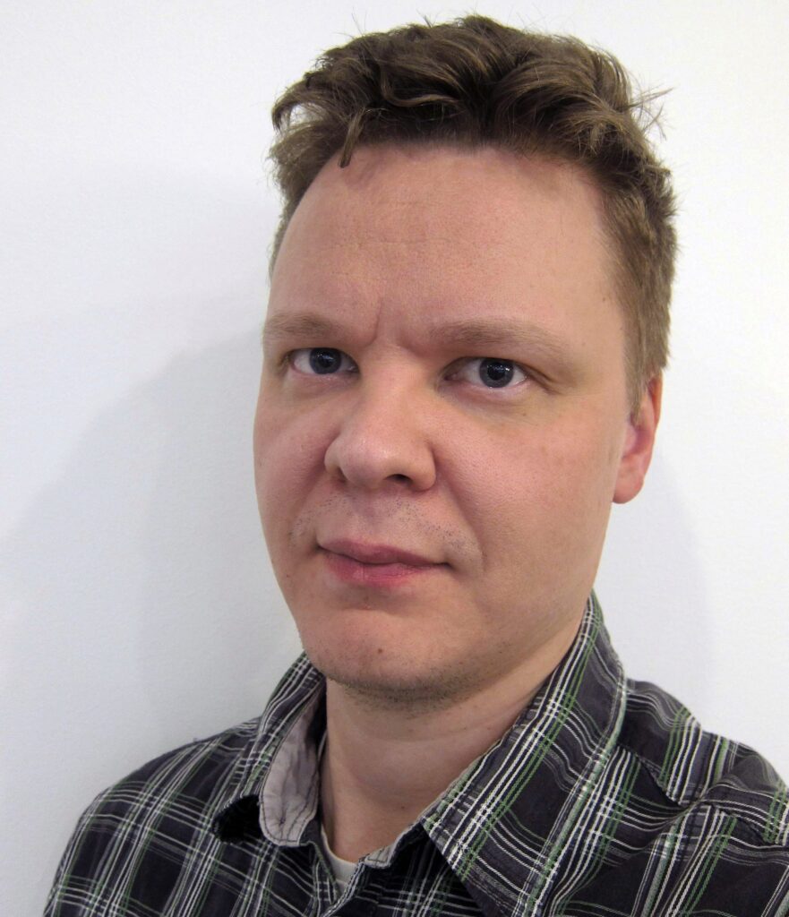 Lyhyet, vaaleanruskeat hiukset ja vaalea iho omaavalla miehellä on yllään ruudullinen paita, ja hän katsoo kameraan neutraalilla ilmeellä valkoista taustaa vasten.