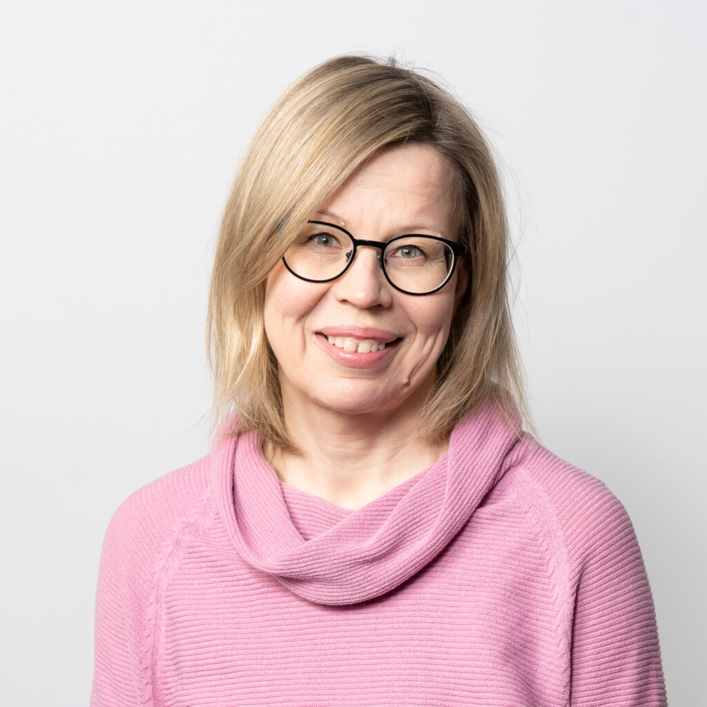 A woman with blonde hair and glasses is smiling. She is wearing a light pink sweater and standing in front of a plain, light background.
