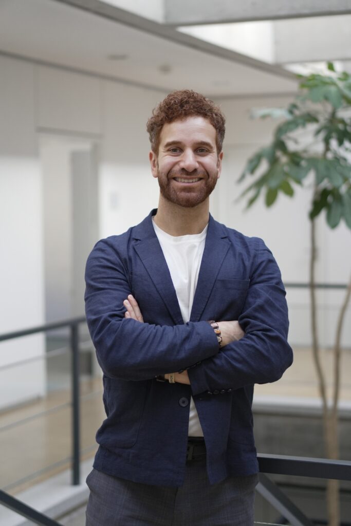A smiling man with curly hair and a beard stands indoors with his arms crossed, wearing a navy blazer and white shirt. A green potted plant and modern office interior are visible in the background.